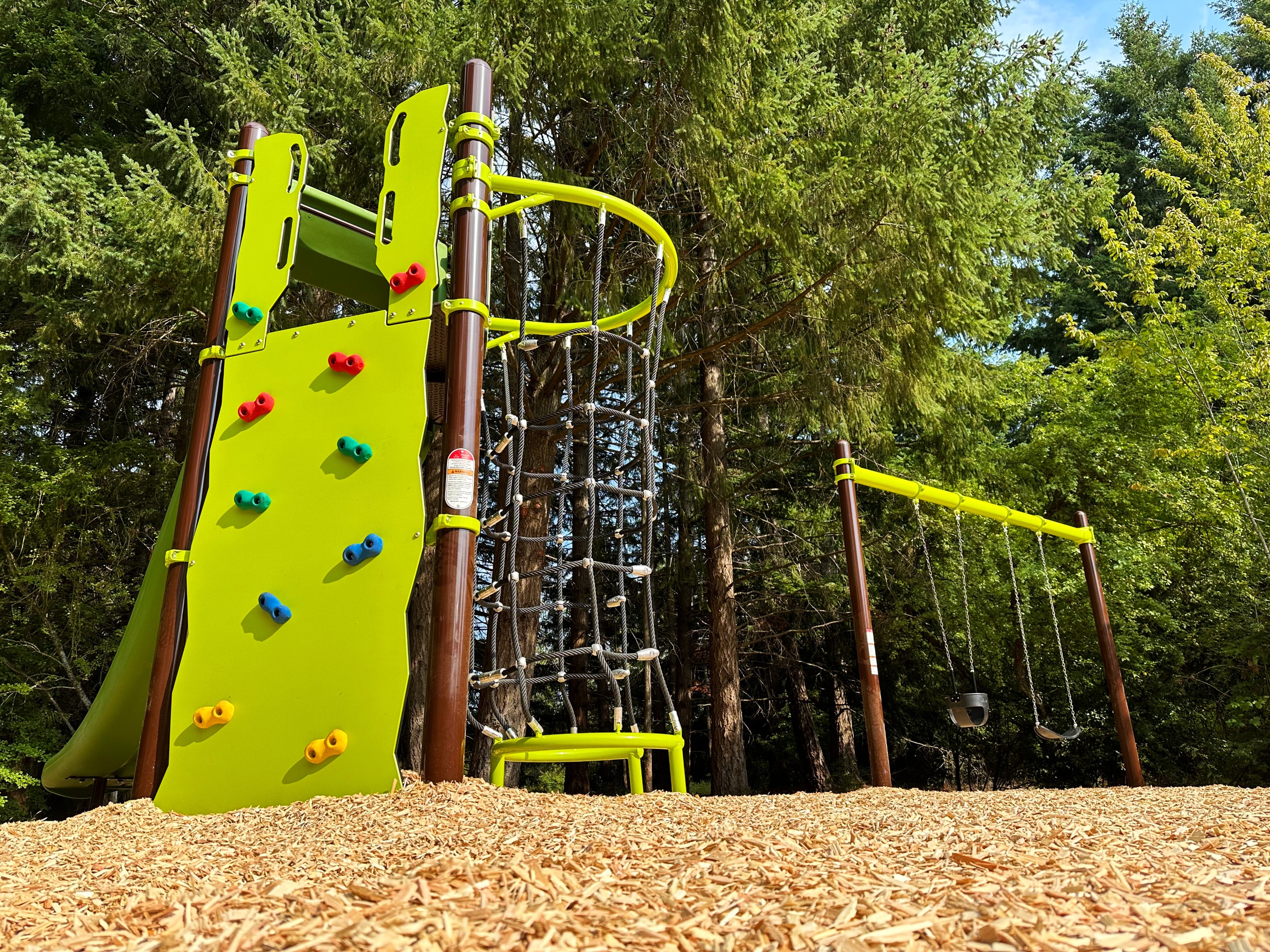 Bright green climbing wall with colorful holds next to a rope ladder and swings in a forested playground covered with wood chips.